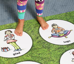 Underbenen på ett barn som står på en matta. På mattan finns illustrationer på barn som utför tecken. Under illustrationerna står ordet för den känsla som tecknet beskriver.