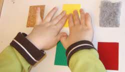 Ett par barnhänder på flexiboard, ett alternativt tangentbord. På Flexiboarden finns ett överlägg med några olika tygbitar med olika struktur som barnen känner på.