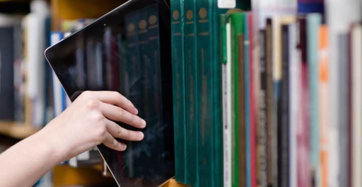 En hand håller en pekplatta halvvägs ut ur en hylla med många tryckta böcker.